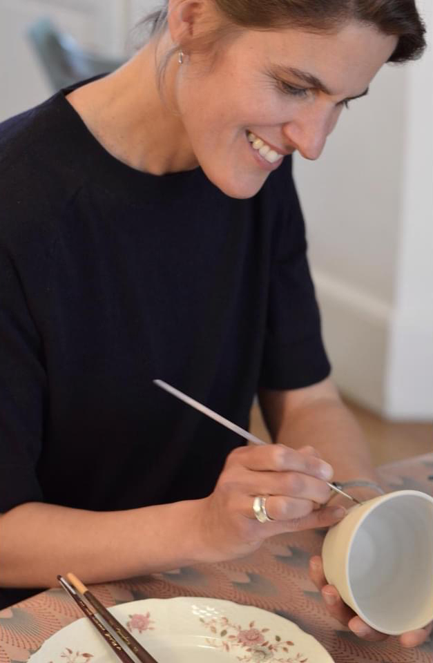 A lady smiling while painting on a ceramic pot.