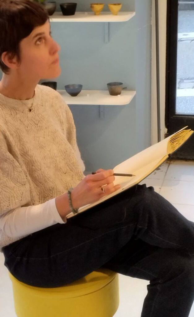 Photograph of the artist, showing her sitting on a yellow seat and drawing.