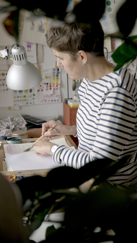 Photograph of Marine drawing at her desk, partly hidden by plants in the foreground.