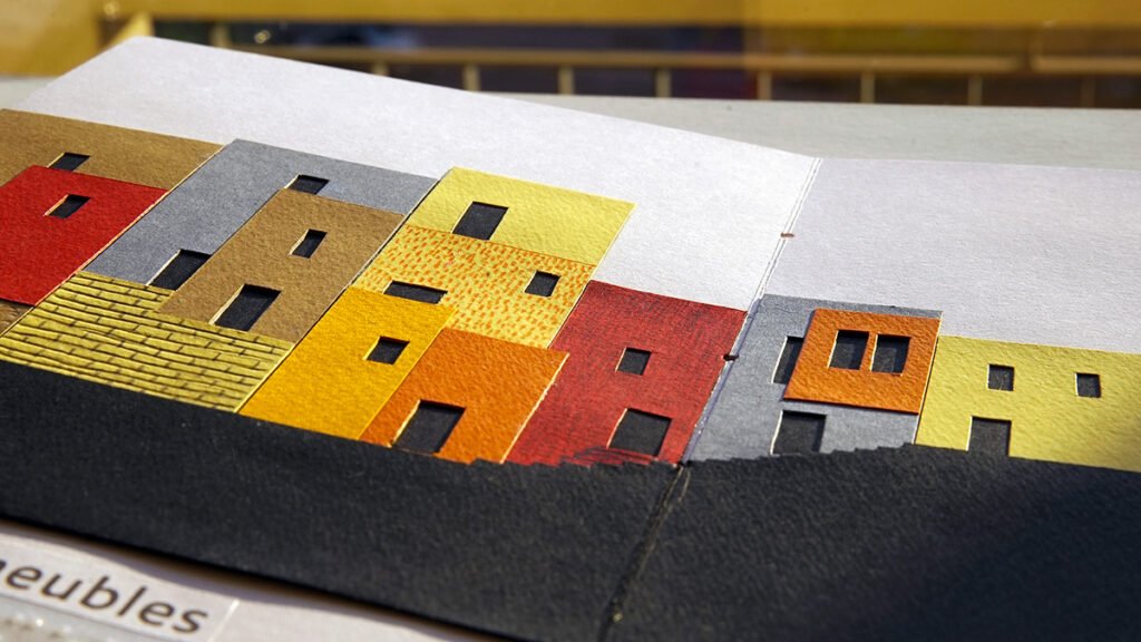 Photographie d'un livre tactile ouvert. On y voit une série d'immeubles de différentes couleurs et dimensions avec de petites fenêtres qui y sont découpées.