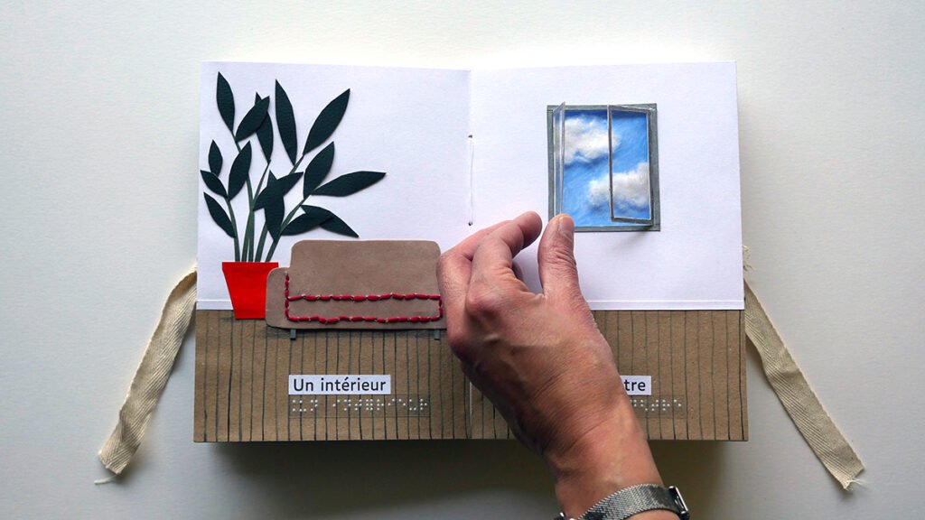 Photographie d'un livre tactile ouvert. Une main ouvre une fen^tre. A gauche on voit un canapé marron et une grande plante verte dans un pot rouge.