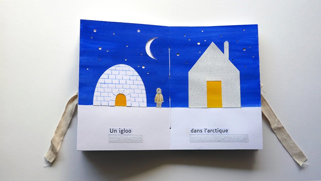 Photographie d'un livre tactile ouvert. Un igloo et une maison, blancs tous les deux, se font face, sur un ciel bleu étoilé.