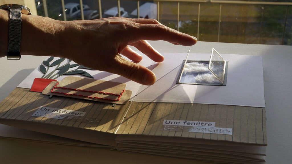 Photographie d'un livre tactile ouvert. Une main s'approche d'une fenêtre pour l'ouvrir.